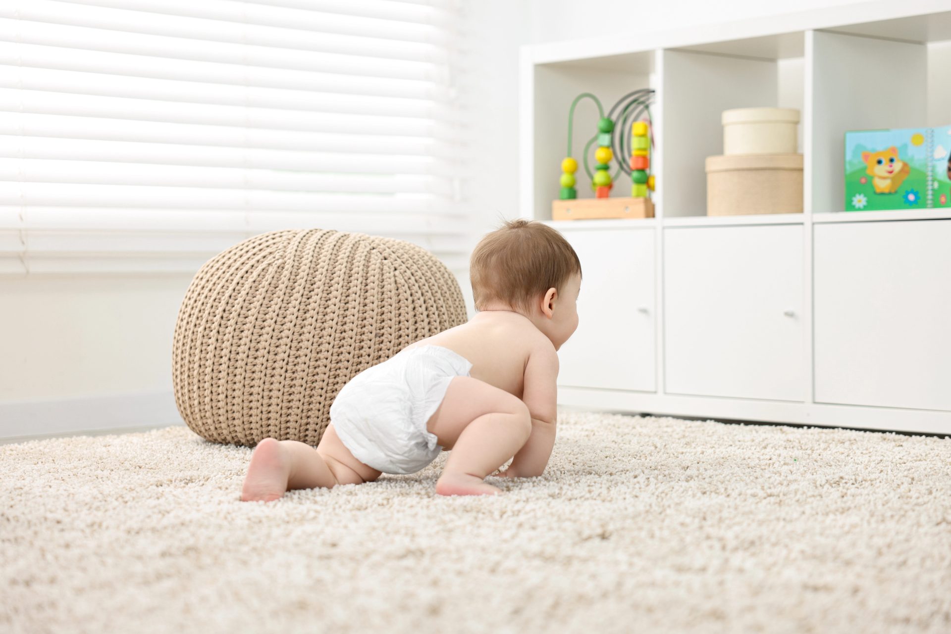 Baby crawling on carpet near white storage unit with toys.