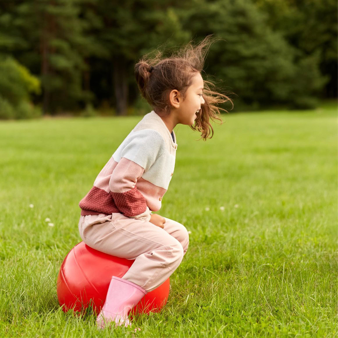 Young girl enjoying outdoor play on a red exercise ball in a green park.