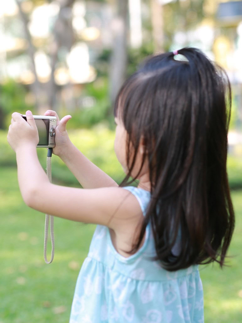 girl-taking-pictures-on-camera-in-the-garden Young girl with dark hair taking a photo with a camera outdoors.