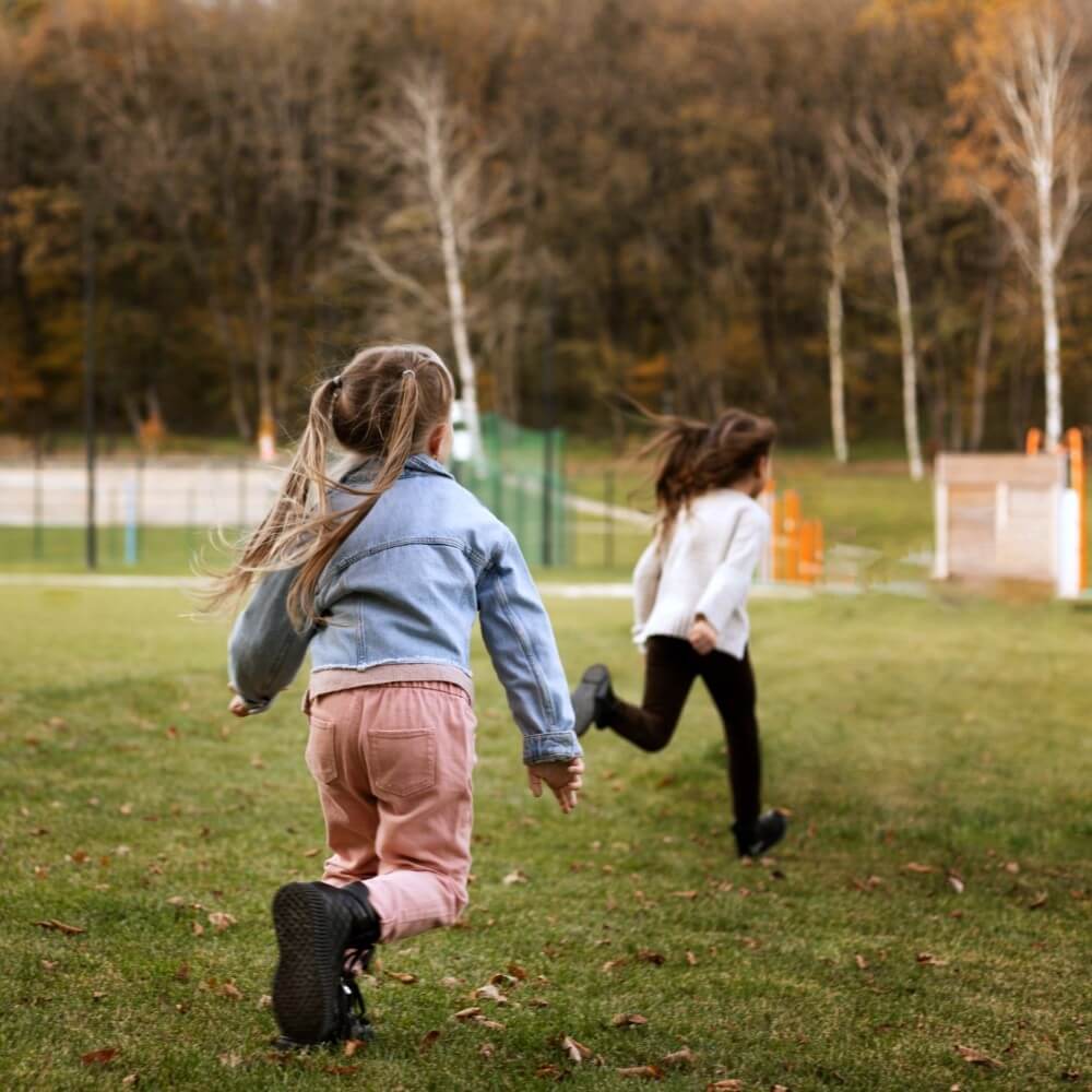 Two children running on a grassy field during autumn with trees in the background.