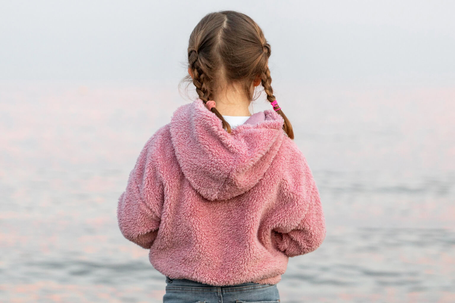 Girl with braided hair wearing pink hoodie facing water.