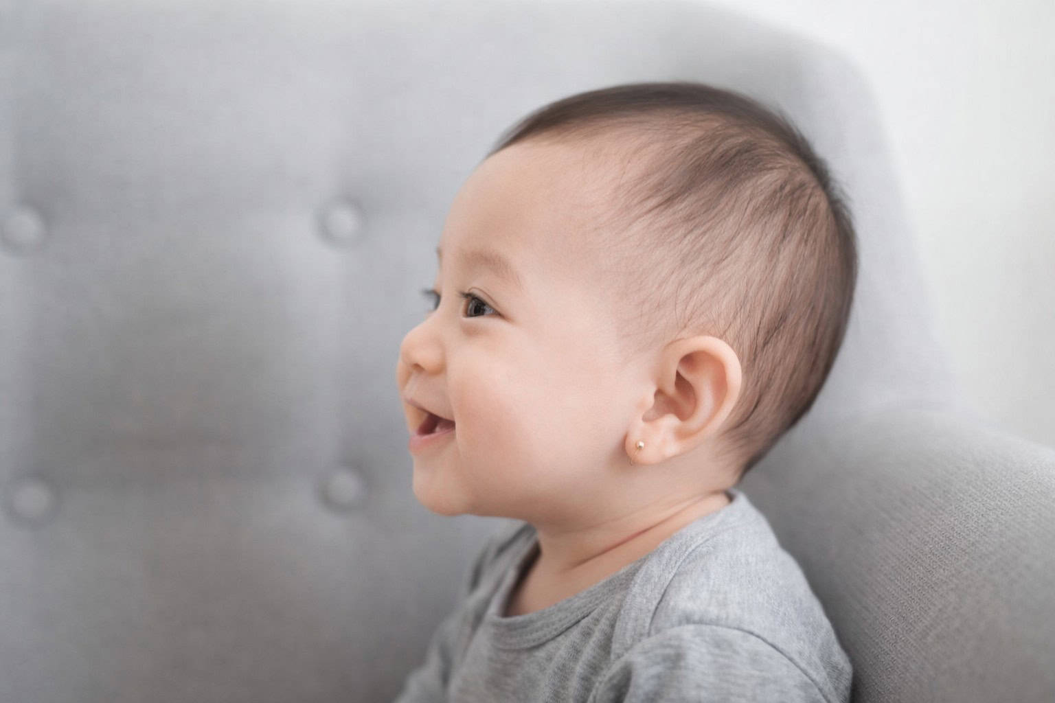 Smiling baby sitting on a gray couch, looking to the side.