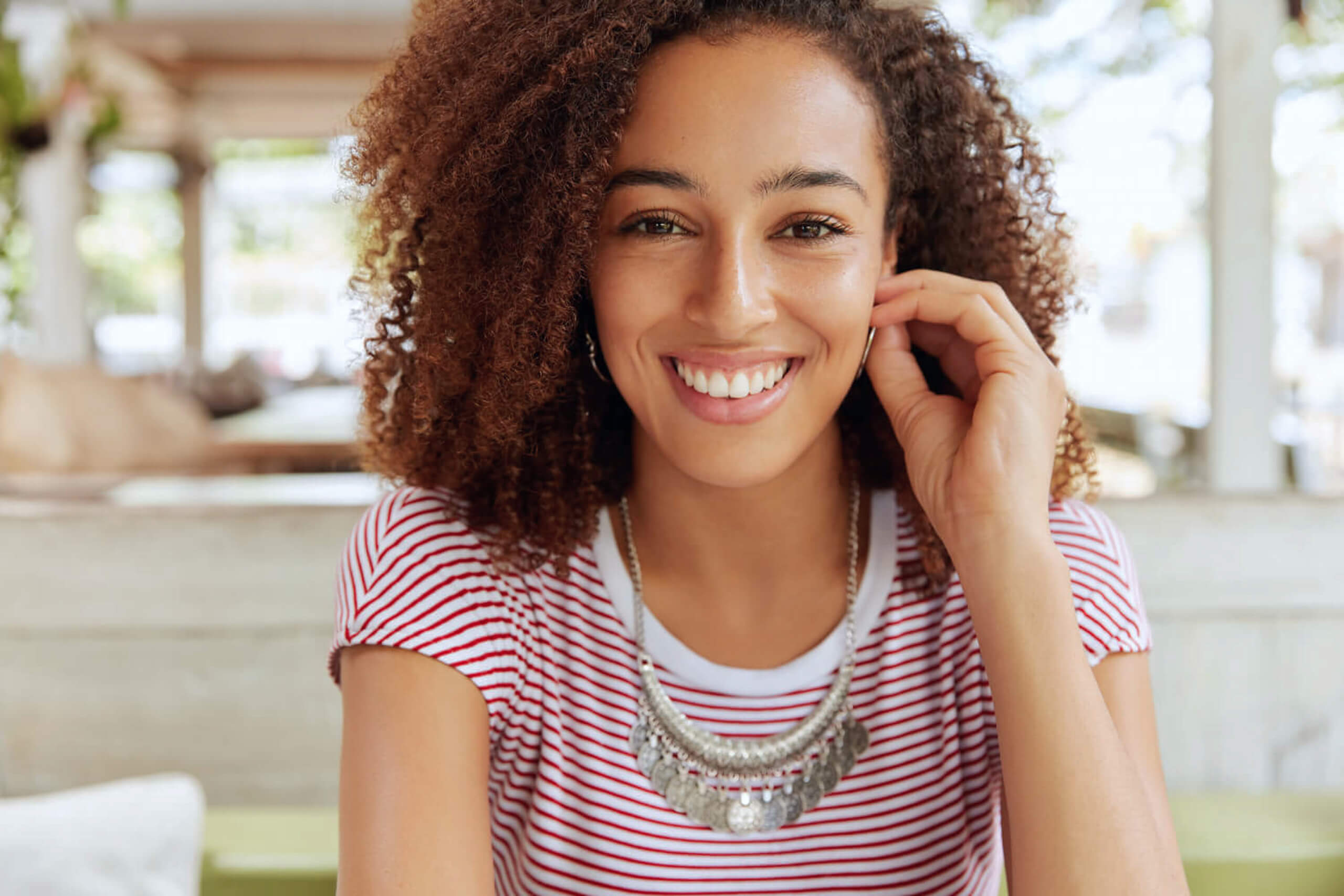 Smiling woman with curly hair in a casual striped shirt at a cafe.