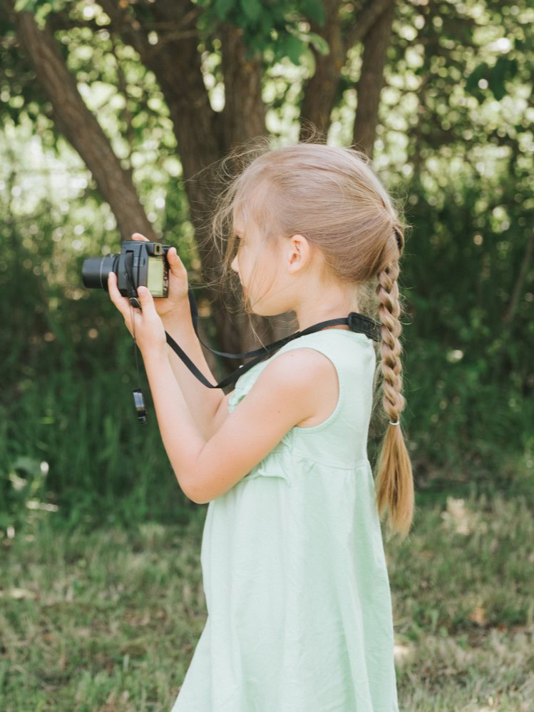 A-happy-little-kid-girl-photographs Young girl with a camera in a green outdoor setting.