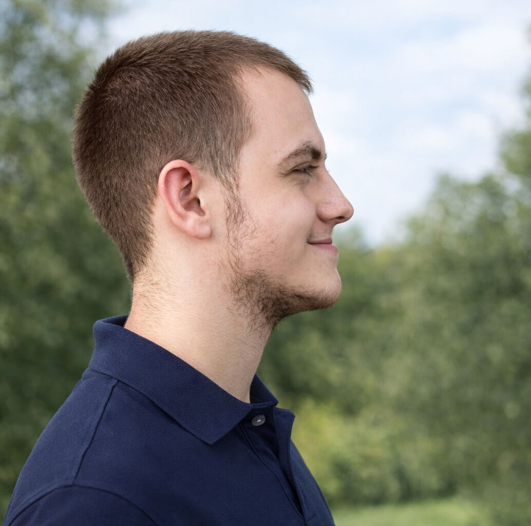Profile of a young man with short hair outdoors, smiling in natural light.