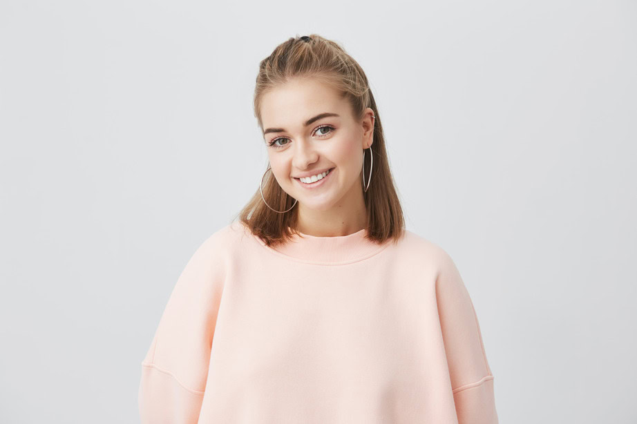 Smiling young woman with light brown hair and hoop earrings in a pink sweatshirt.
