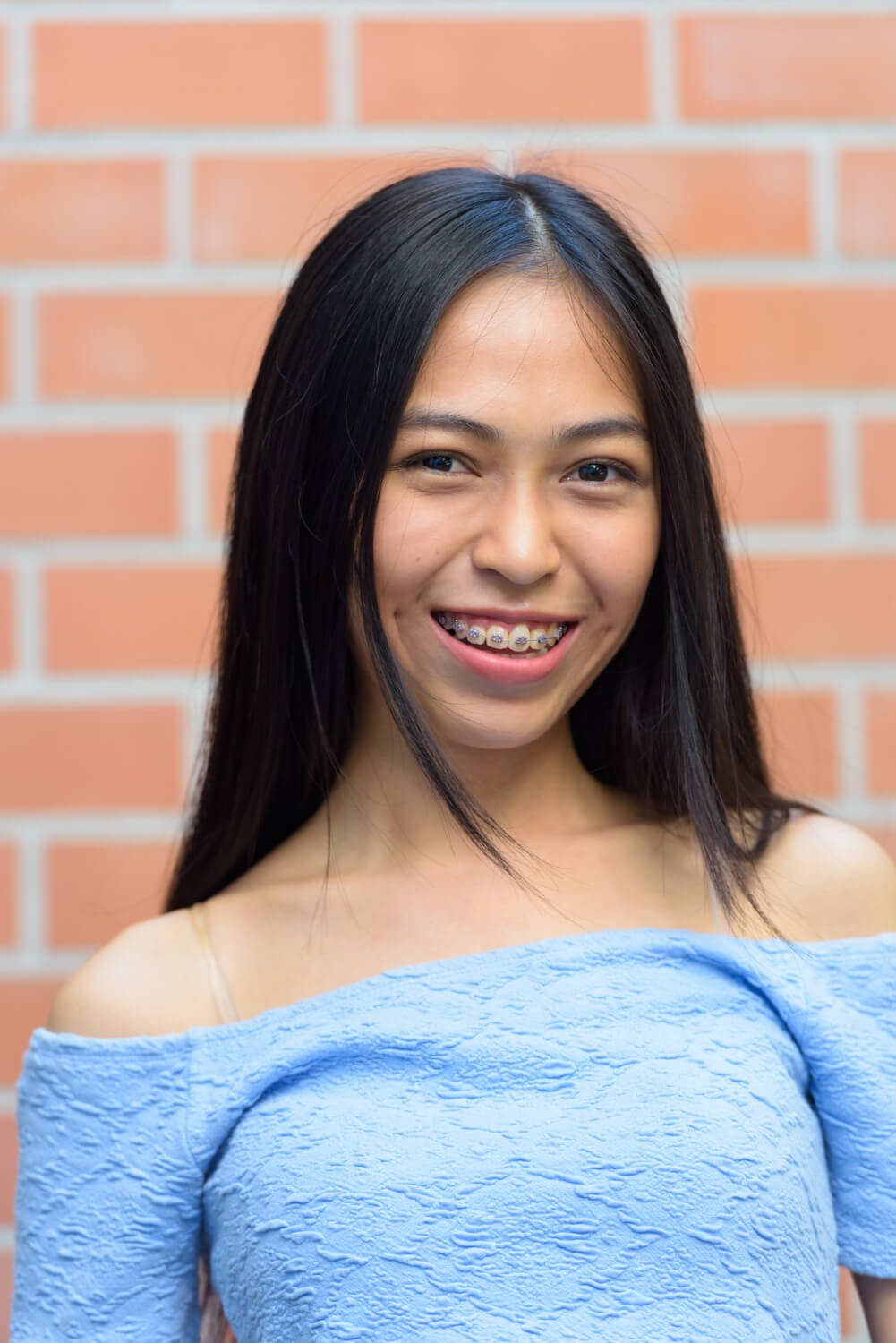 Smiling young woman with braces and long black hair in front of brick wall.