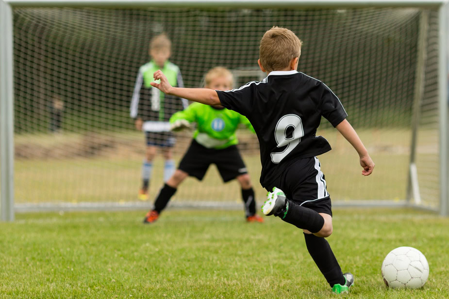 Young boy in black soccer uniform kicking a ball towards goal.